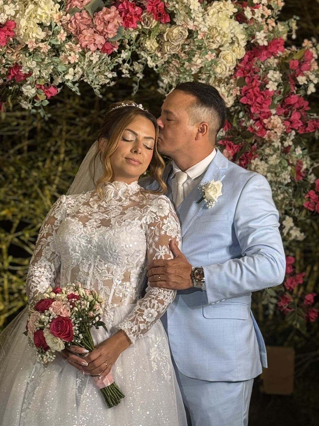 Noivo beija a testa da noiva diante de arco de rosas